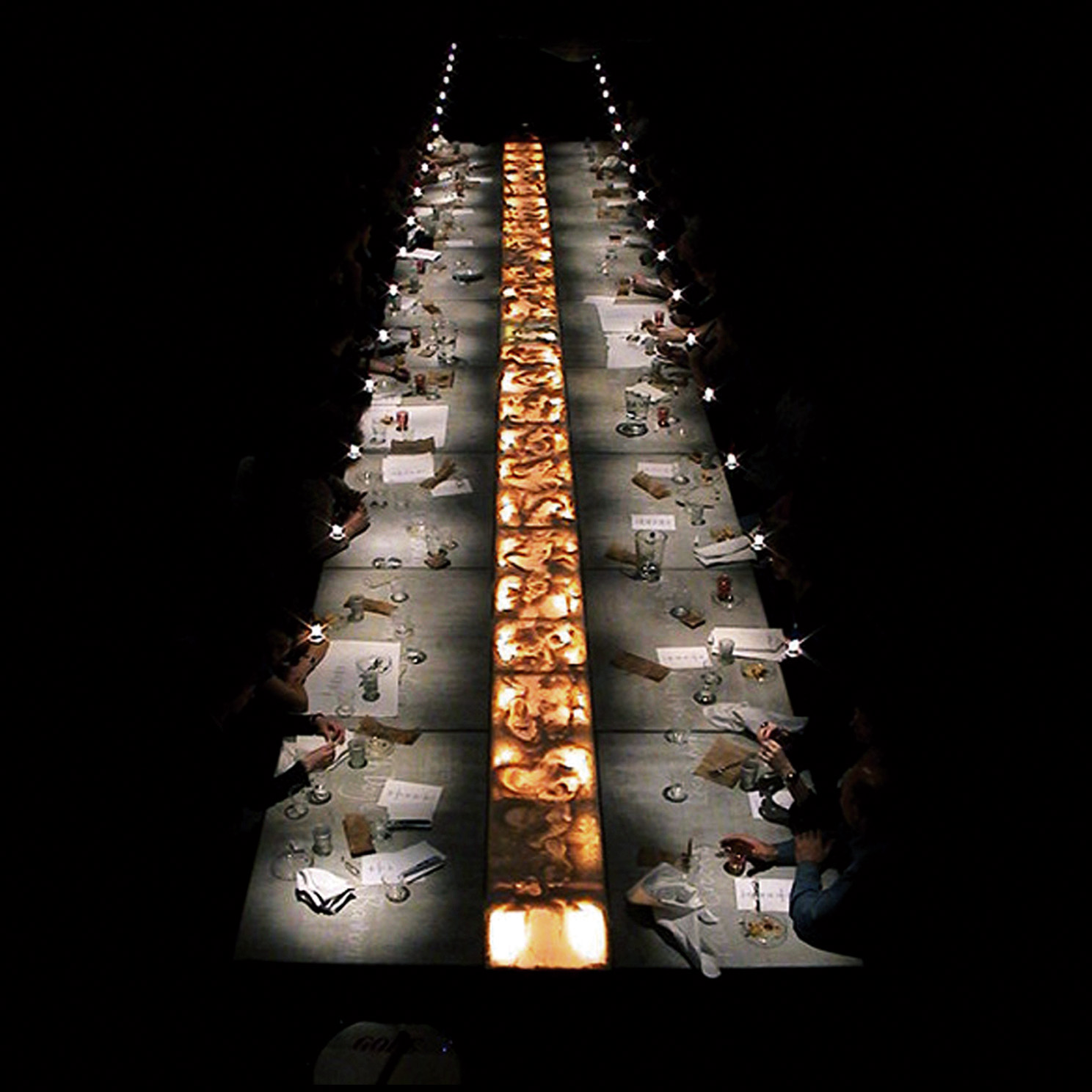 An aerial shot of a long table with place settings lit centrally, with diners in the dark, can just see their hands