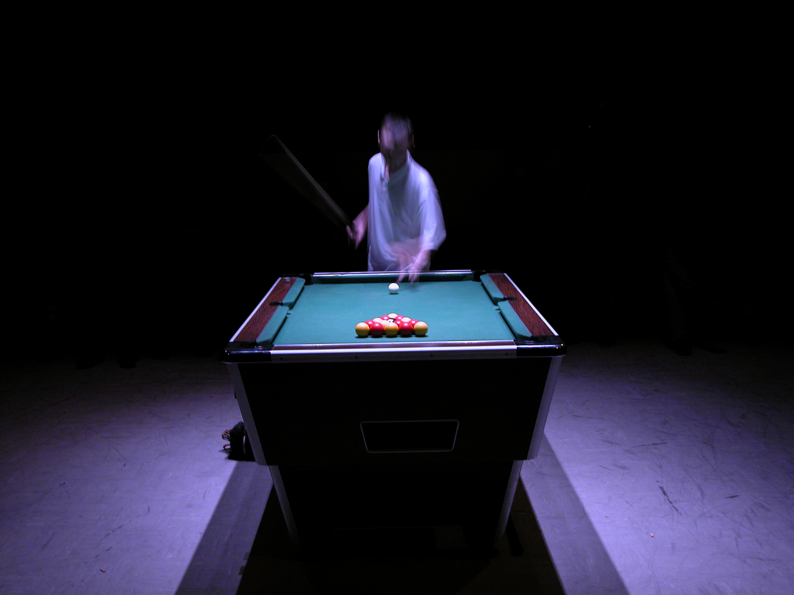 A pool table in sharp focus with a blurred person in a white shirt holding a cue