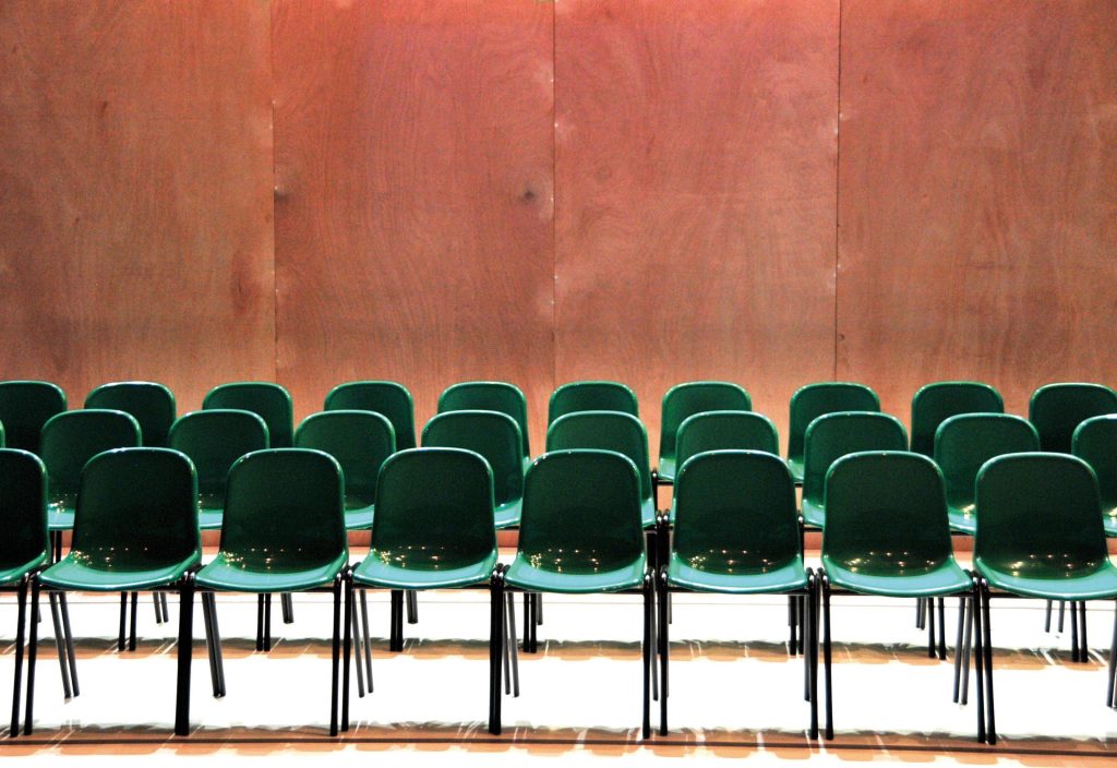 Wooden panelled wall at the back with 3 rows of green plastic chairs in the foreground