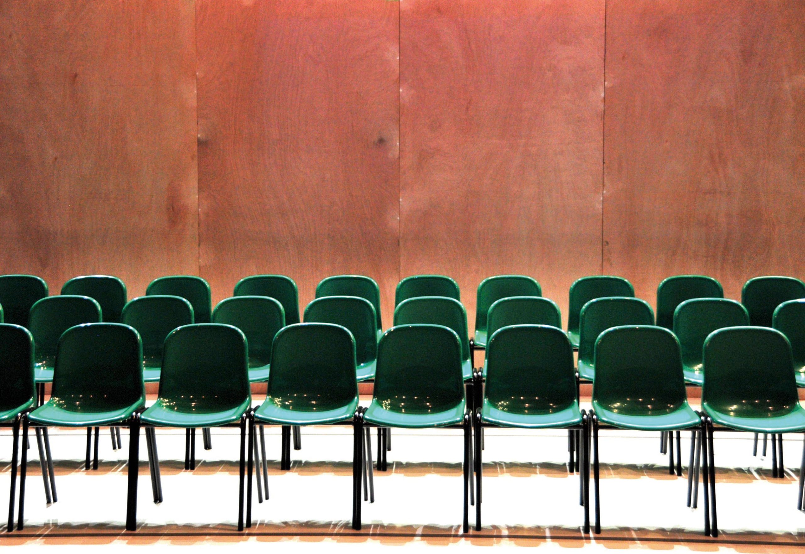 Wooden panelled wall at the back with 3 rows of green plastic chairs in the foreground
