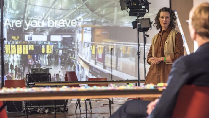 A woman in a brown dress stands in front of a screen with a projected question using 'Are you brave?'. The screen also displays a live feed of a railway station. In the foreground a person is sat on an orange chair with their back to the camera. They are looking at each other.