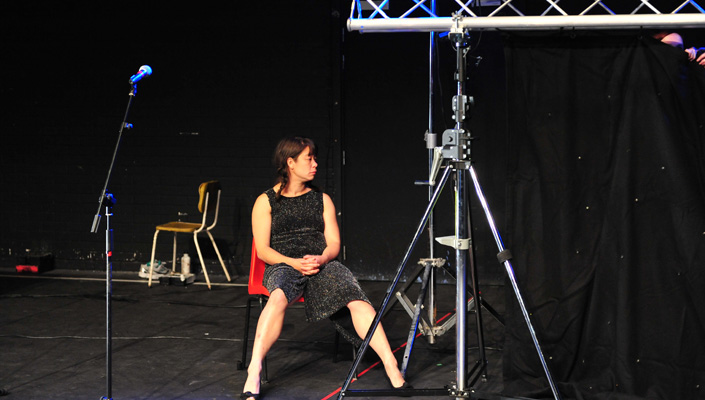 Dark haired woman sits on an orange chair in a black box theatre. Looking away from the camera. There is a microphone on a stand to the left of her. She is wearing a glittery black dress.