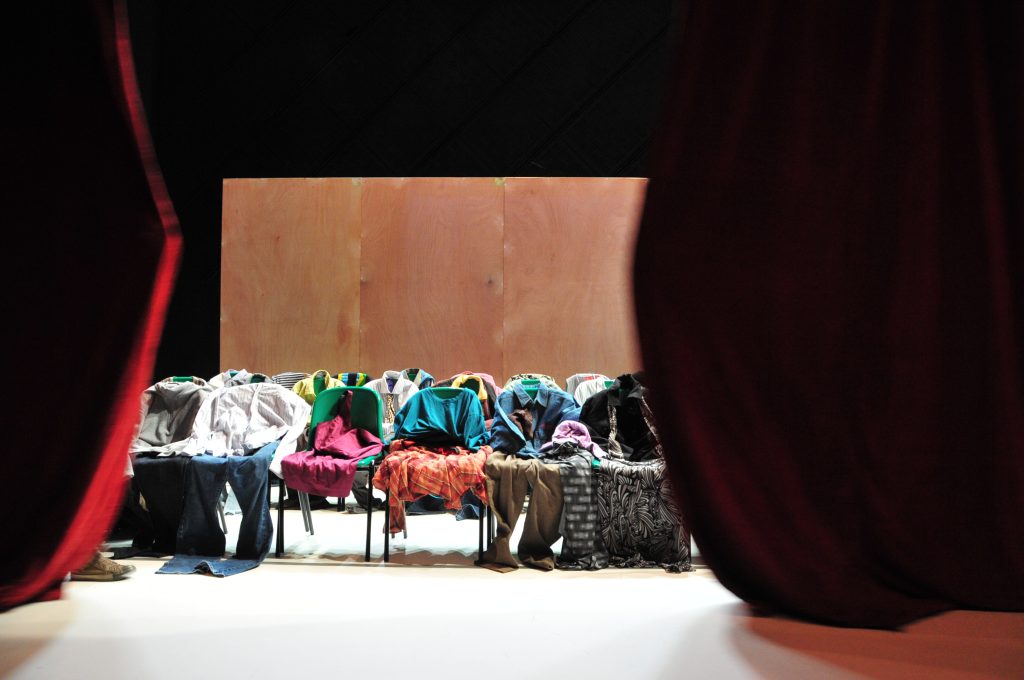 Clothes on chairs visible on stage behind a red curtain