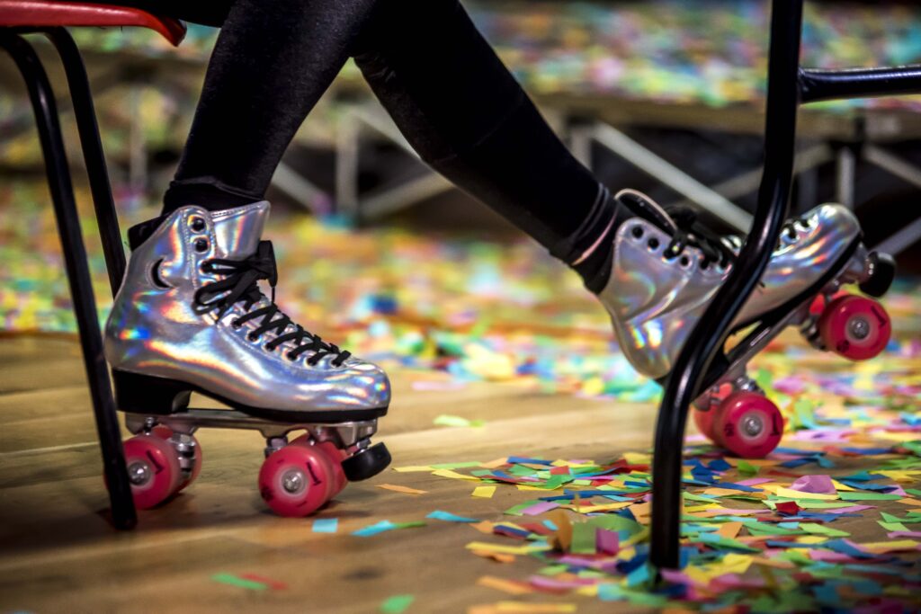 The ankles of a skater who is sitting at a table, wearing irridesent rollerskates, with red wheels. The floor is covered in confetti.