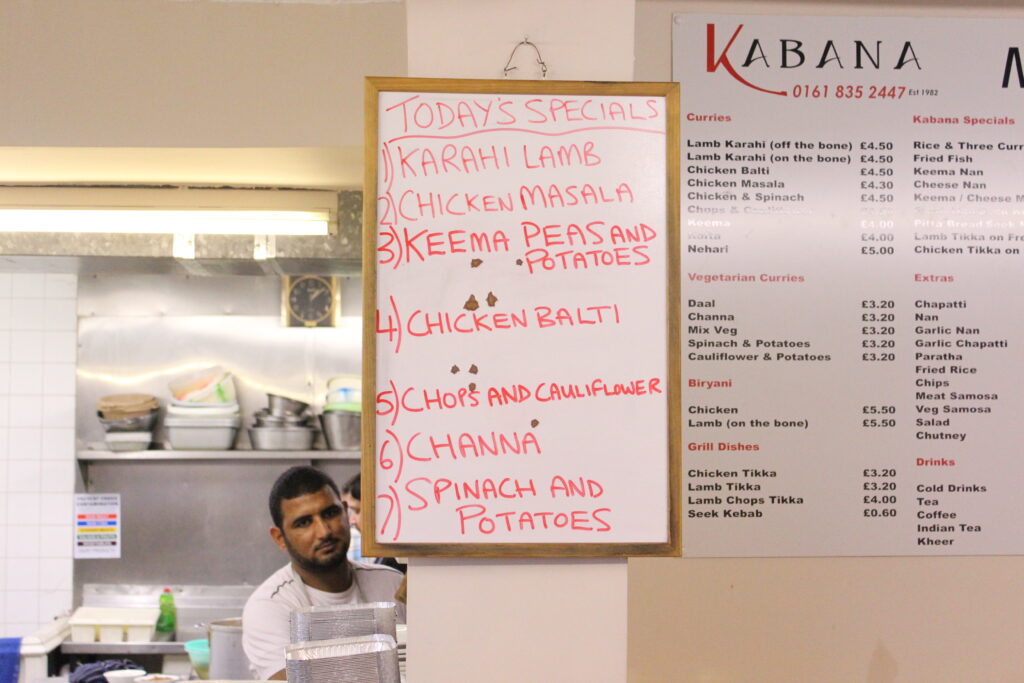 A whiteboard is in the centre of the picture with a list of special dishes offered by the cafe - to the right a full menu of food option - to the left of the whiteboard we see the kitchen and one of the chefs looking directly at us