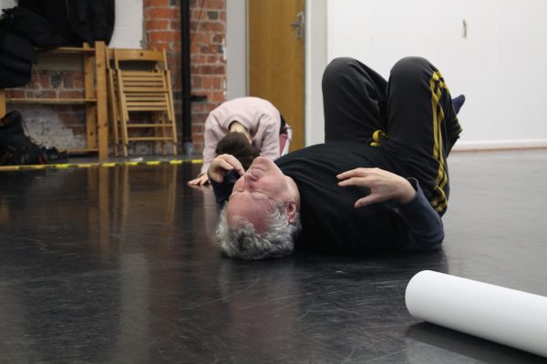 Man wearing all black on dance floor of studio. Laid on his back with knees raised, eyes closed. Woman in pink jumper kneeling in background, head on knees arms stretched out front.